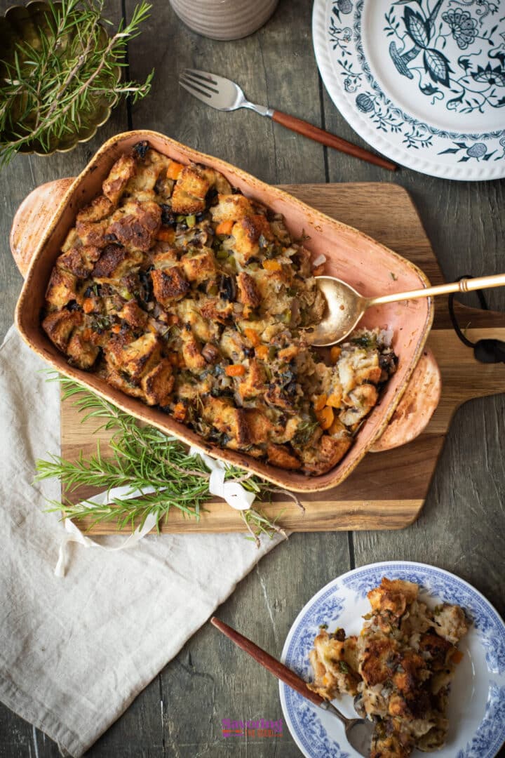 A baking dish filled with bread stuffing sits on a wooden board, with a serving spoon and fresh herbs nearby; a portion is served on a blue-and-white plate.