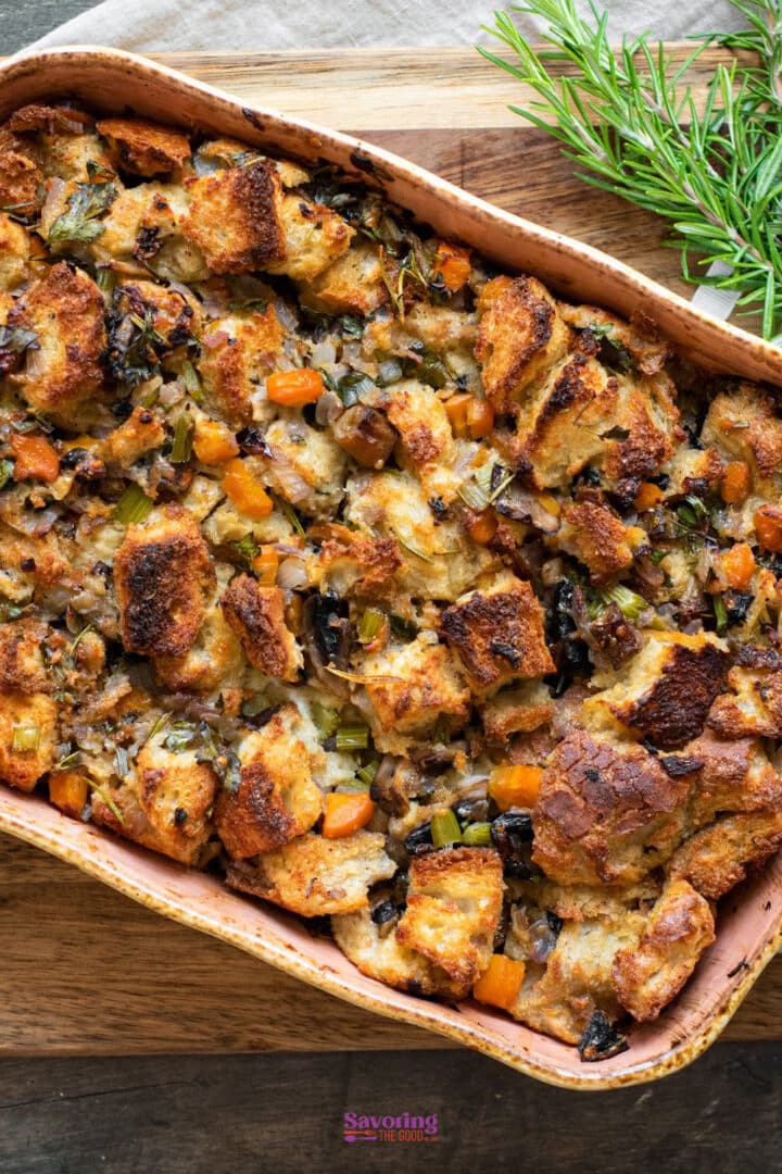 A baked casserole dish filled with bread stuffing mixed with chopped vegetables and herbs, placed on a wooden surface next to fresh rosemary.
