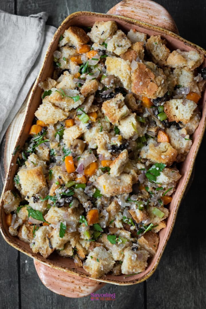 Rectangular baking dish filled with cubed bread stuffing mixed with vegetables and herbs, sitting on a dark wooden surface next to a beige linen napkin.