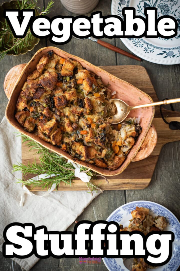 A ceramic baking dish with vegetable stuffing sits on a wooden board, a serving spoon inside. Fresh herbs and a plate with a portion of stuffing are nearby.