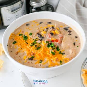 A bowl of creamy soup reminiscent of Crockpot cream cheese chicken chili with tender chicken, corn, black beans, diced tomatoes, shredded cheese, and a parsley garnish, placed on a white surface.