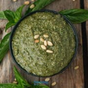 A bowl of green pesto topped with pine nuts, surrounded by fresh basil leaves on a wooden surface.