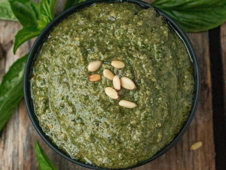 A bowl of green pesto topped with pine nuts, surrounded by fresh basil leaves on a wooden surface.