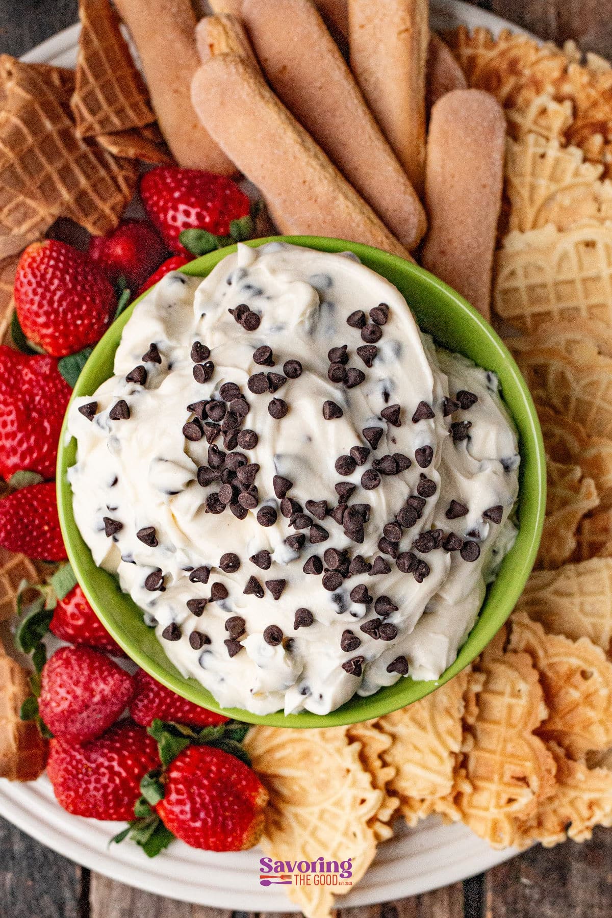 A bowl of cannoli dip topped with chocolate chips, surrounded by strawberries, ladyfingers, and waffle cookies.