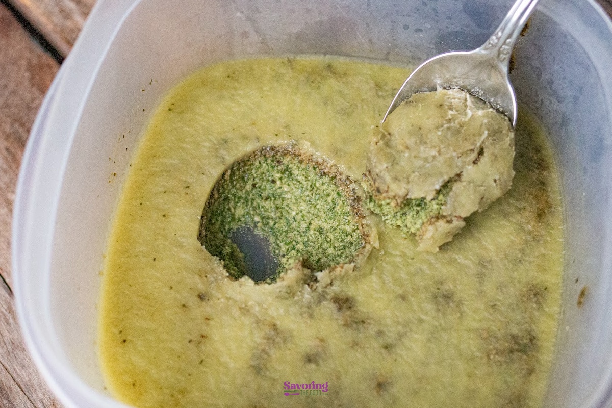 A metal spoon lifts a scoop of thick, pale green purée from a plastic container, revealing a bright green, textured interior.