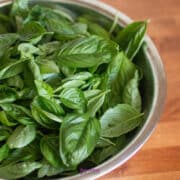 A metal bowl filled with fresh green basil leaves sits on a wooden surface.