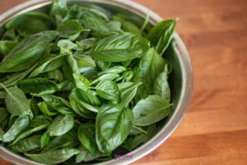 A metal bowl filled with fresh green basil leaves sits on a wooden surface.