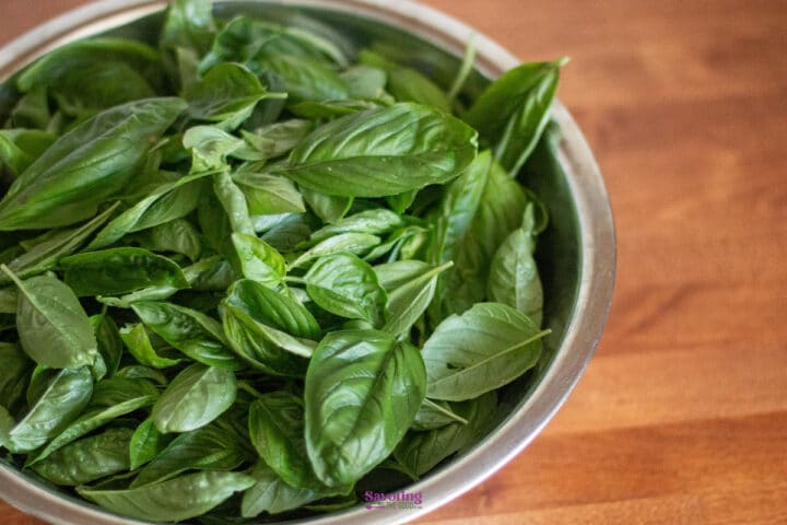 A metal bowl filled with fresh green basil leaves sits on a wooden surface.