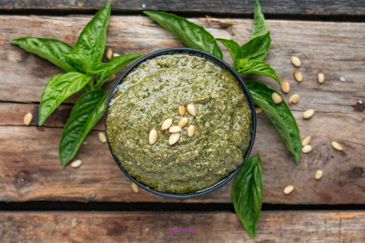 A bowl of green pesto garnished with pine nuts, surrounded by fresh basil leaves on a wooden surface.
