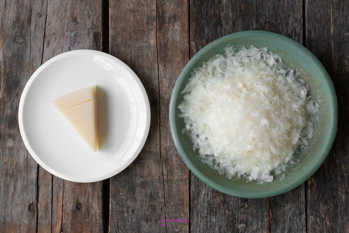 A wedge of parmesan cheese on a white plate next to a bowl of shaved parmesan cheese on a wooden table.