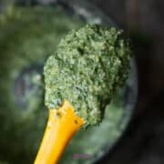 A yellow spatula holding a scoop of blended green pesto, with a food processor bowl in the background.
