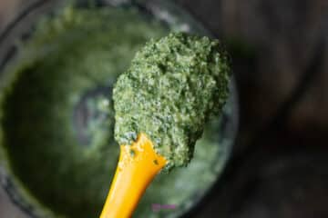A yellow spatula holding a scoop of blended green pesto, with a food processor bowl in the background.