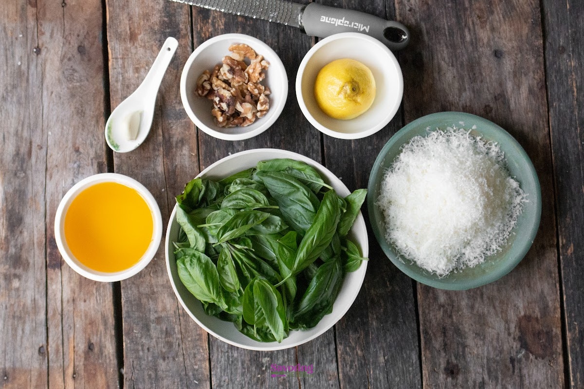 Ingredients for pesto including fresh basil, grated cheese, walnuts, a lemon, olive oil, and a clove of garlic, arranged on a wooden surface with a zester.