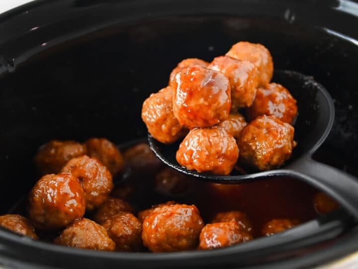 A black slotted spoon lifts several glazed grape jelly meatballs from a slow cooker filled with sauce.
