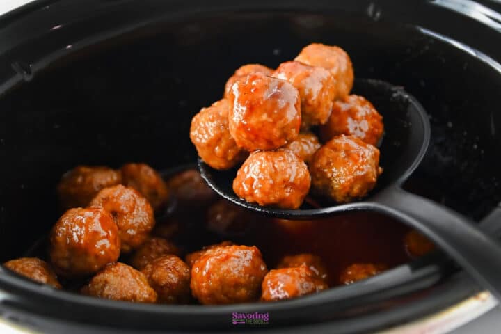 A black slotted spoon lifting glazed grape jelly meatballs from a slow cooker filled with sauce.
