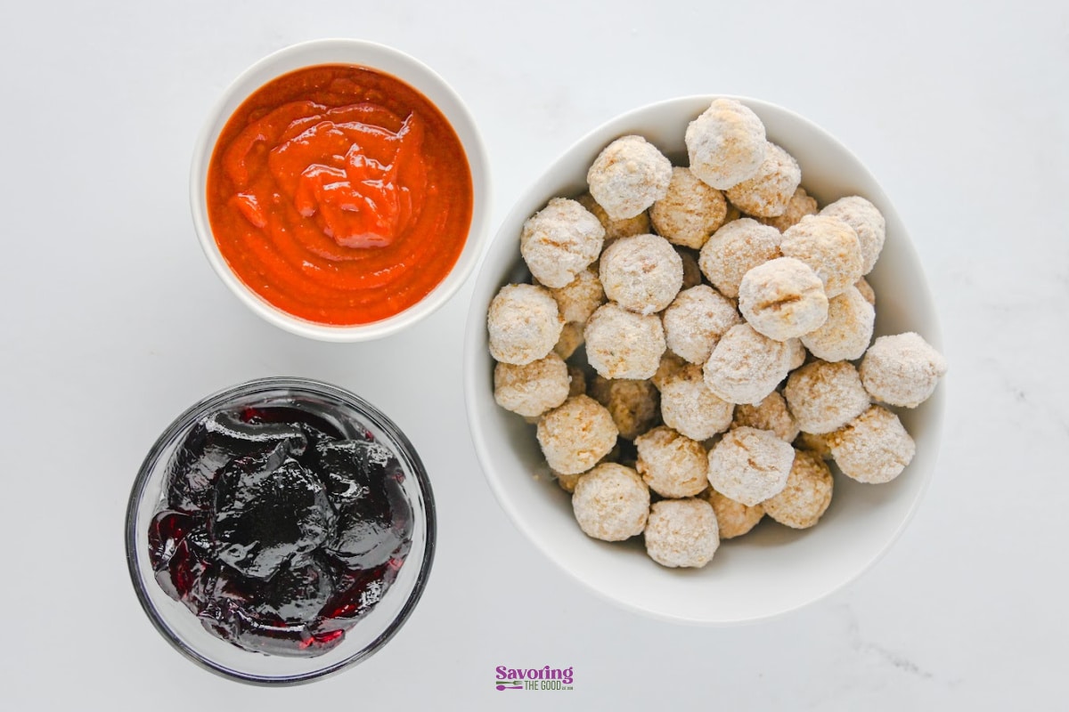 A large bowl of breaded appetizer bites served with a bowl of red marinara sauce and a bowl of dark grape jelly meatballs dipping sauce.