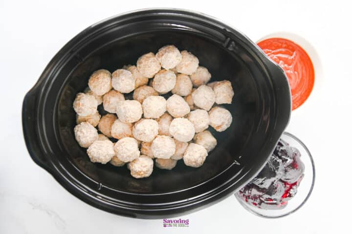 A slow cooker filled with frozen grape jelly meatballs, positioned next to small bowls of red sauce and dark jam.