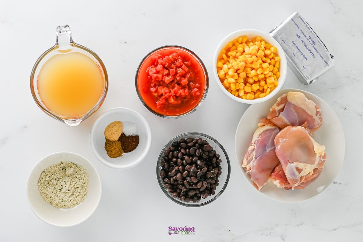 Ingredients for a recipe arranged on a white surface, including chicken thighs, black beans, corn, diced tomatoes, broth, spices, cream cheese, and a dry seasoning mix.
